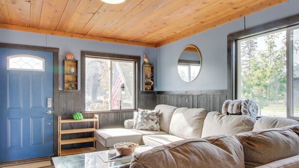A cozy living room with a wooden ceiling, a blue door, a large window, a beige sofa, and a glass coffee table. - Quaint Ironton Getaway w/ Fire Pit, Gas Grill! (Ironton)