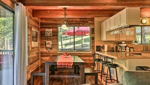 A cozy cabin interior with a wooden dining table, bar stools, a kitchen area with cabinets and a window overlooking a scenic landscape. - Spacious Groveland Cabin w/ Wraparound Deck! (Groveland)