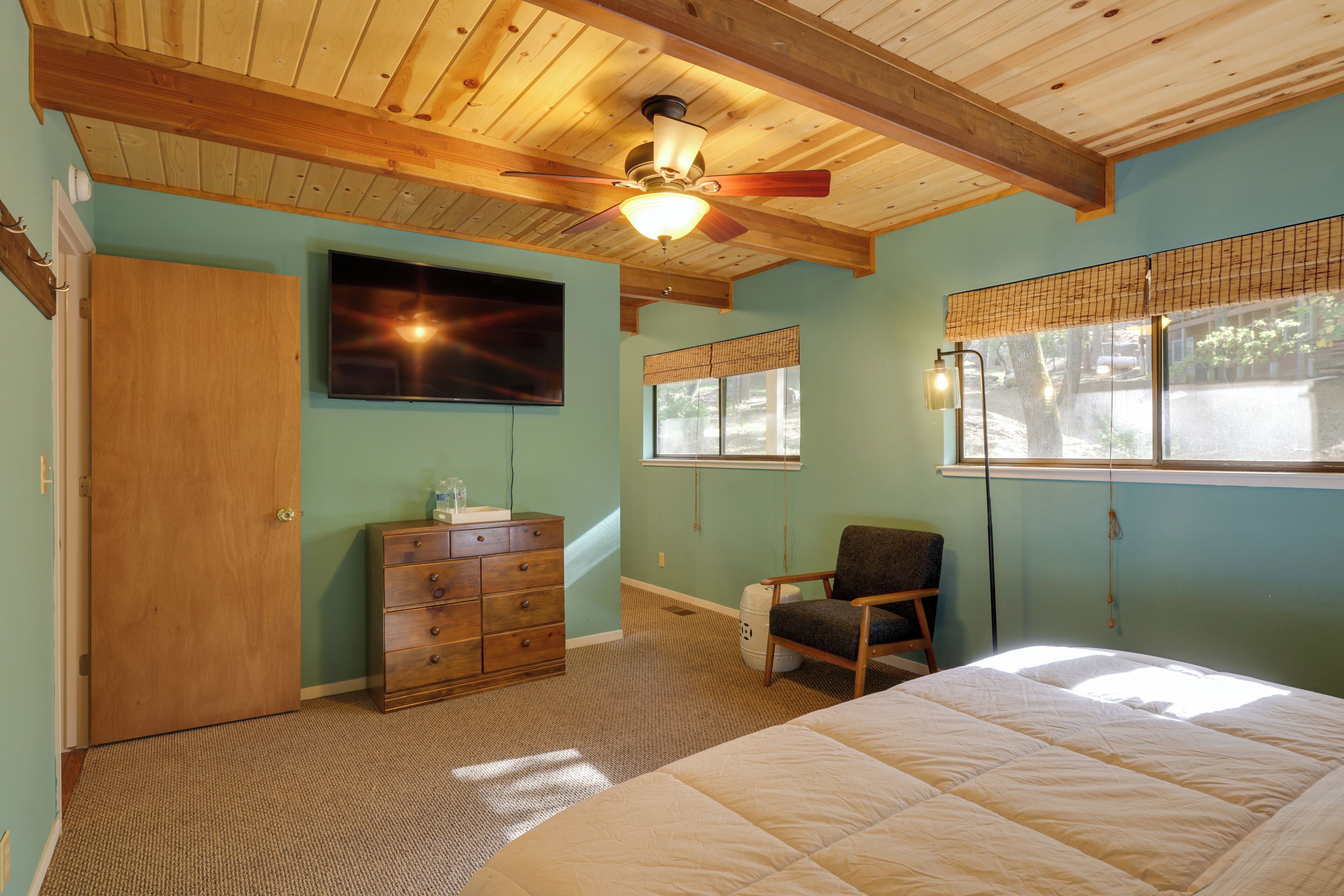 A bedroom with a wooden ceiling, a bed, a dresser, a chair, and a window with blinds.