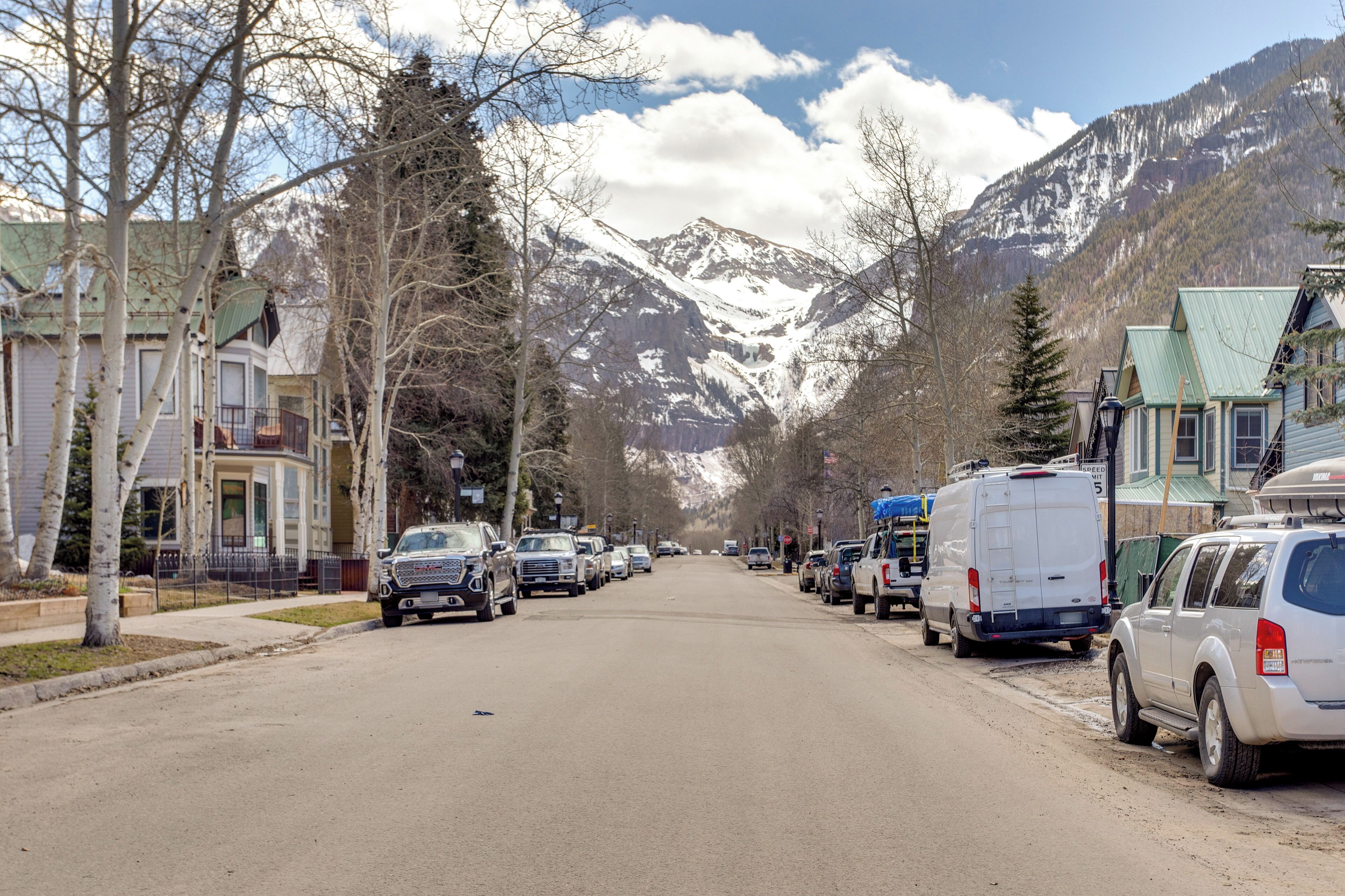 A street lined with parked cars and houses, with mountains in the background.