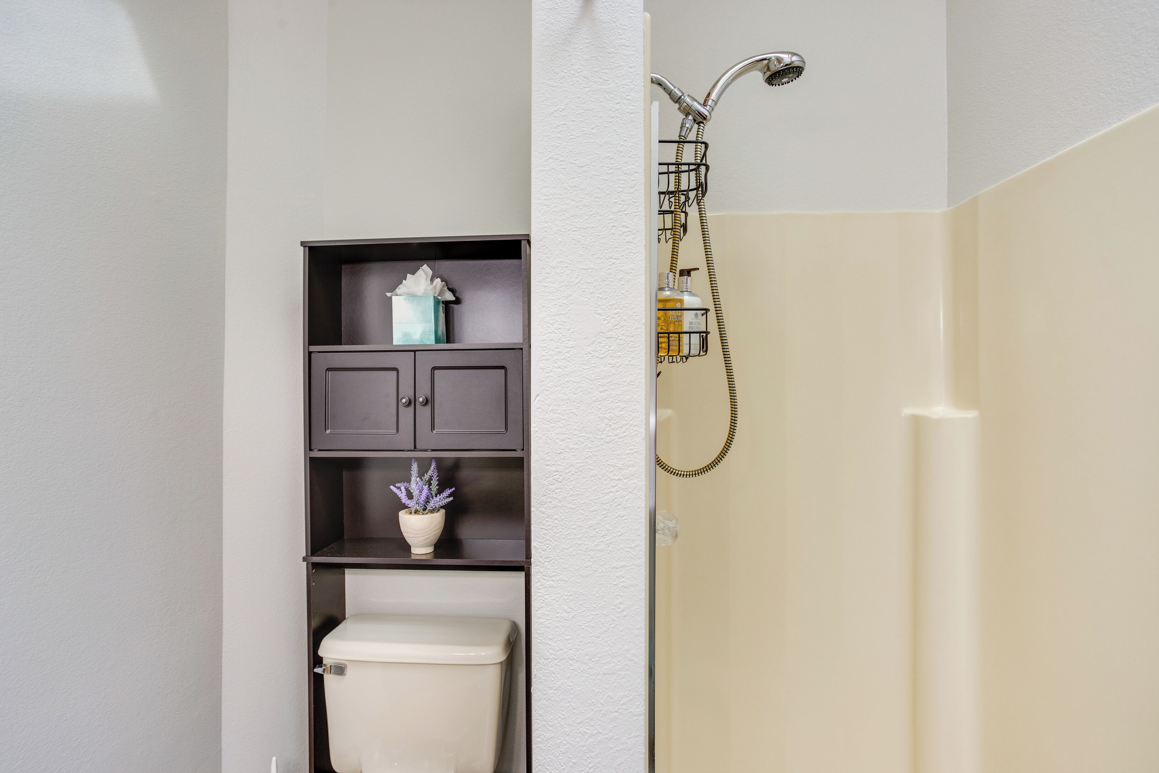 A bathroom with a toilet, a shelf with a tissue box and a potted plant, and a shower area with a towel rack.