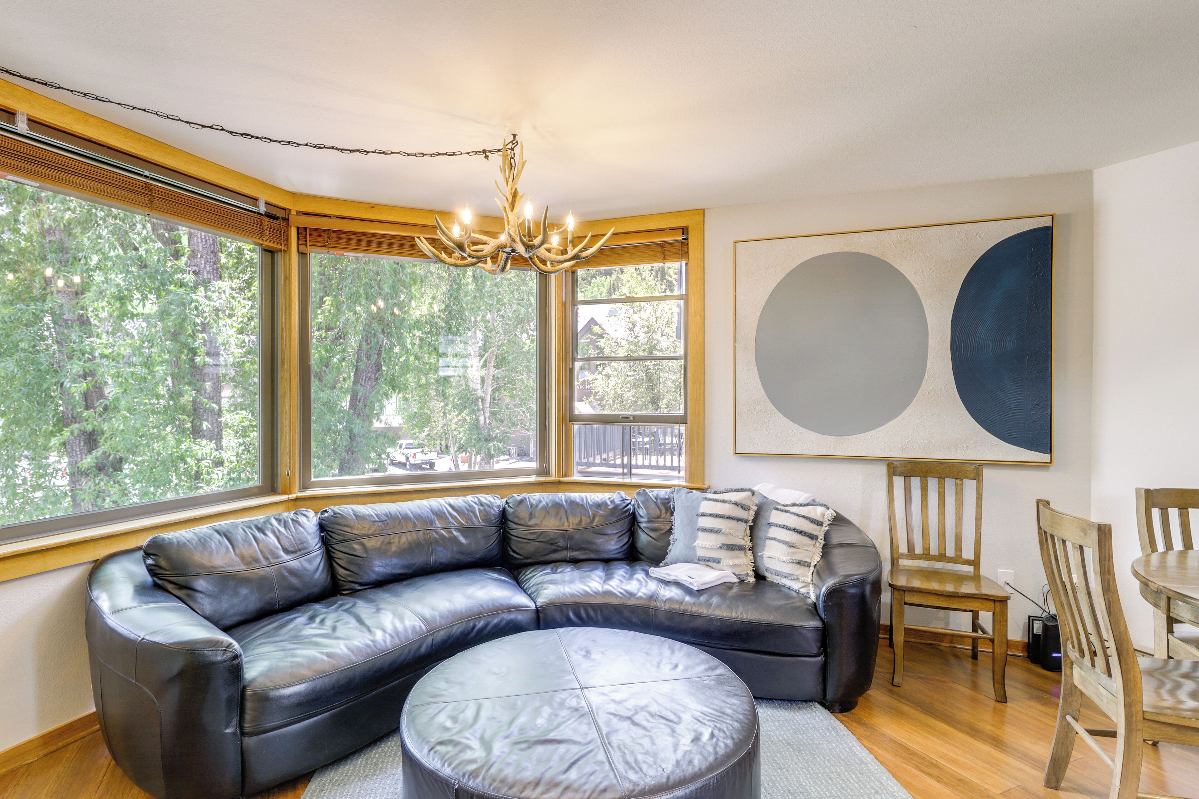 Una sala de estar moderna con un sofá seccional de cuero, un otoman redondo, una mesa de comedor de madera y sillas. Amplias ventanas ofrecen vistas al exterior.