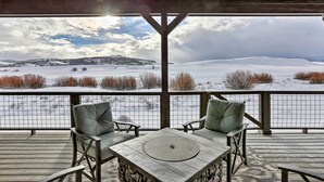 A covered outdoor seating area with two cushioned chairs and a table, overlooking a snowy landscape with distant hills. - Granby Golf Retreat w/ Ski Trails, Mountain View! (Granby)