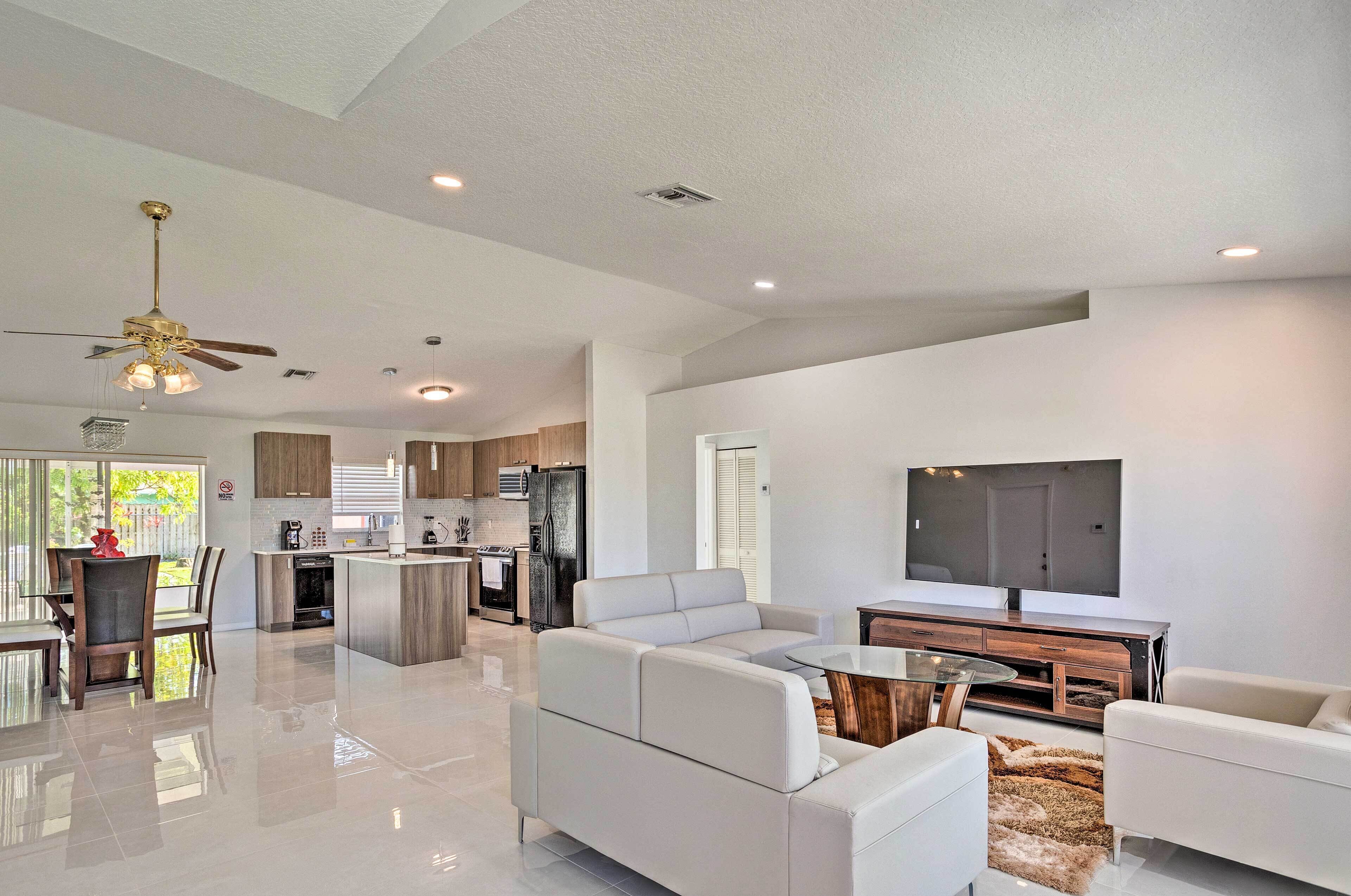 A modern living room with a large TV, white sofas, a glass coffee table, and a ceiling fan.