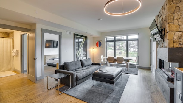 Un salon moderne avec un canapé gris, une table basse et une cheminée. Un téléviseur est fixé au mur et une fenêtre laisse entrer la lumière naturelle.