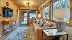A cozy cabin interior with a wooden ceiling, a sofa with cushions, a table with a lamp, and a television on the wall. - Leavenworth Cabin w/ Private Hot Tub! (Leavenworth)