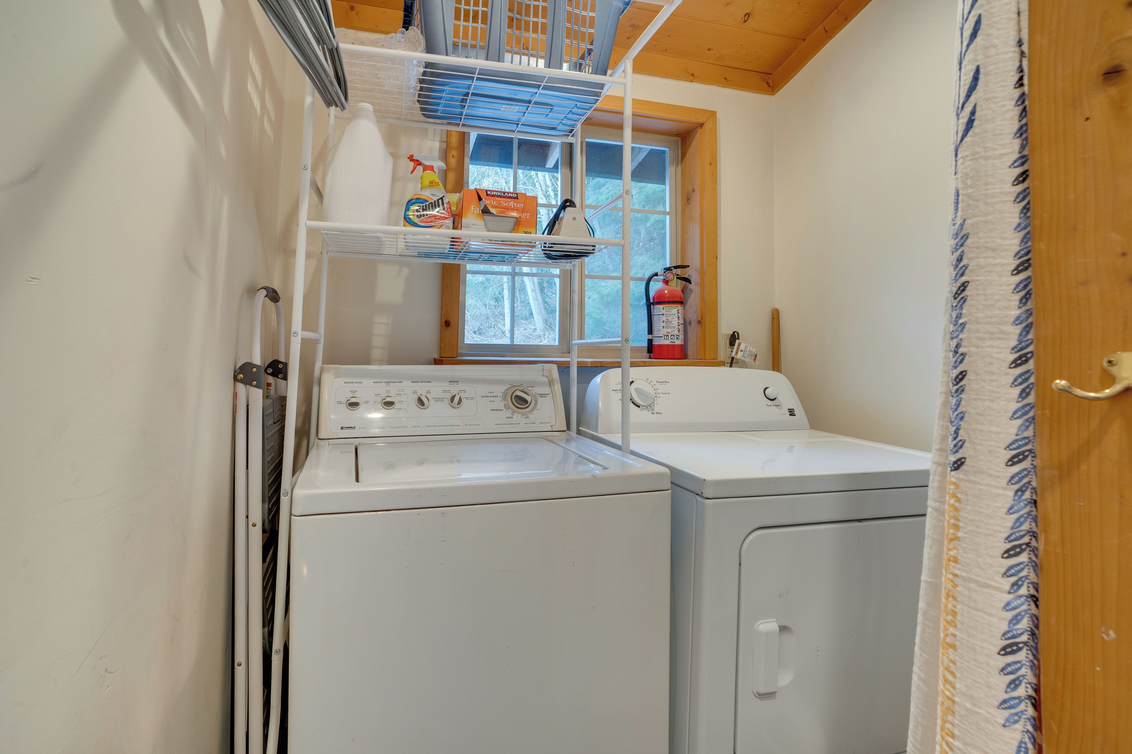 A laundry room with two white washing machines, a shelf with cleaning supplies, a window with a view of trees, and a fire extinguisher.