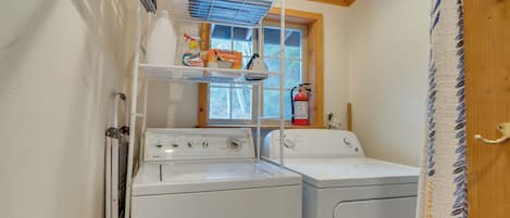A laundry room with two white washing machines, a shelf with cleaning supplies, a window with a view of trees, and a fire extinguisher.