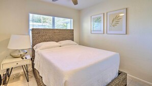 A neatly arranged bedroom with a bed, a bedside table with a lamp, a ceiling fan, and two framed botanical prints on the wall.