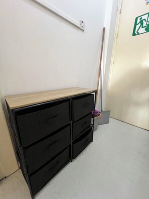 Ein Zimmer mit einer schwarzen Aufbewahrungsbox an der Wand, einem an die Wand gelehnten Besen und einer Tür mit einem Notausgangsschild.