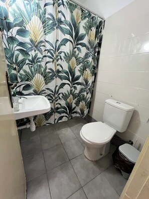 Ein Badezimmer mit Toilette, Waschbecken und einer Duschtasse, deren Vorhang ein tropisches Blattmuster zeigt.