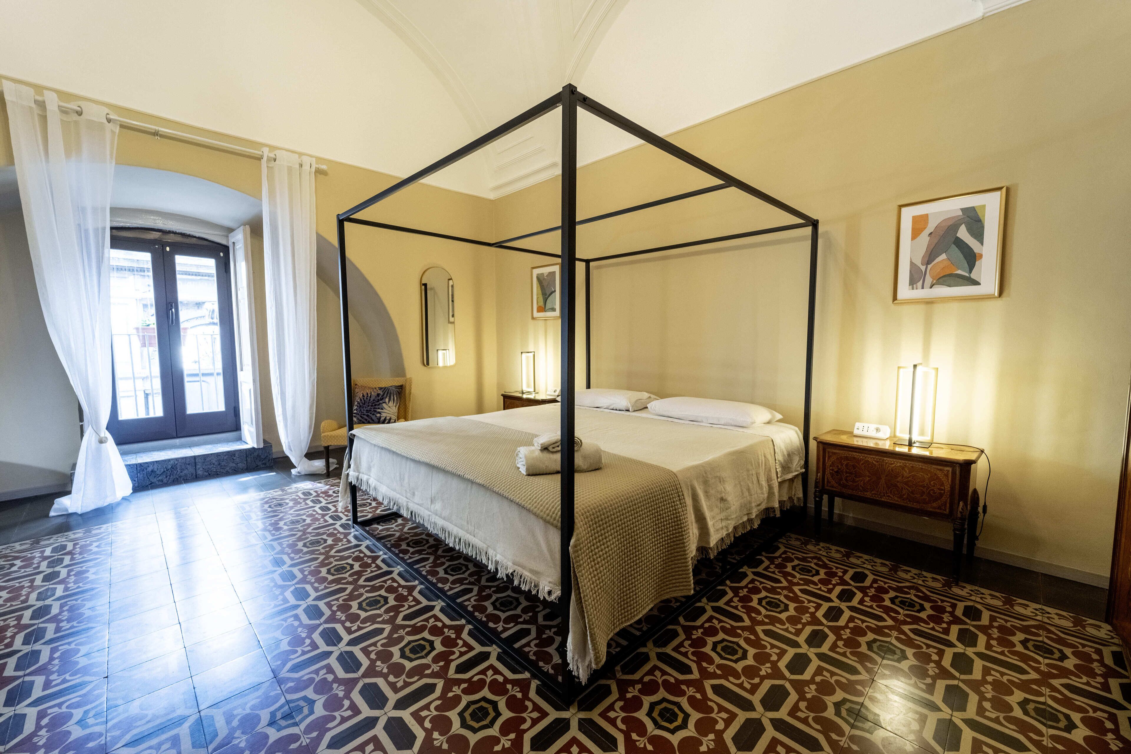 A four-poster bed with a white bedspread, a bedside table with a lamp, and a patterned floor.