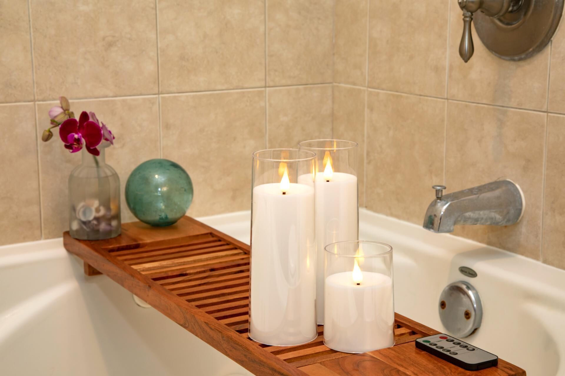 A bathroom with a wooden tray holding three white candles, a teal vase with flowers, and a remote control.
