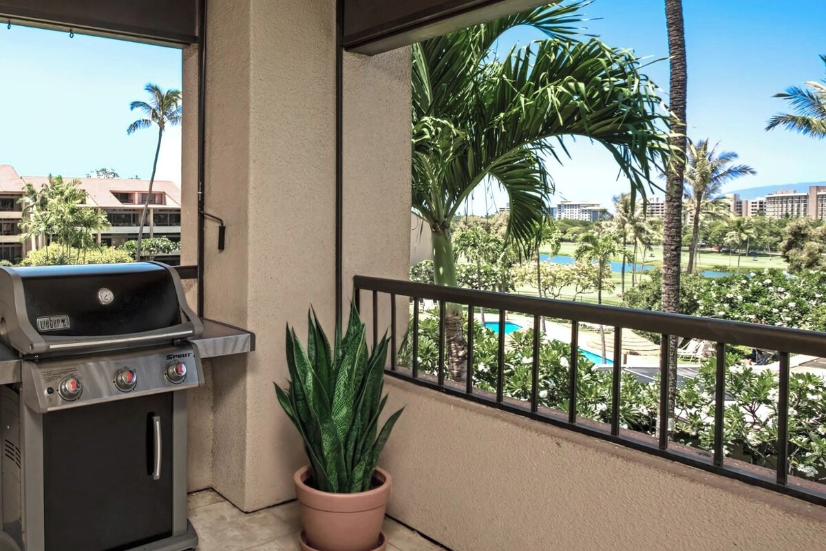 A balcony with a grill and a potted plant, overlooking a pool and palm trees.