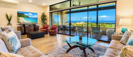 A spacious living room with a large TV, comfortable seating, a glass coffee table, and a view of the ocean through large windows.