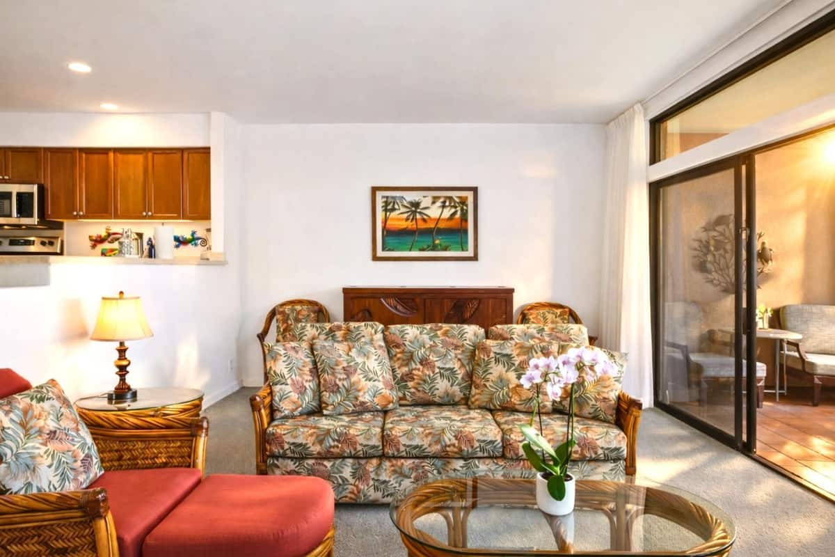 Ein Wohnzimmer mit einer Couch, zwei Sesseln, einem Glastisch und einem Bild einer tropischen Landschaft.
