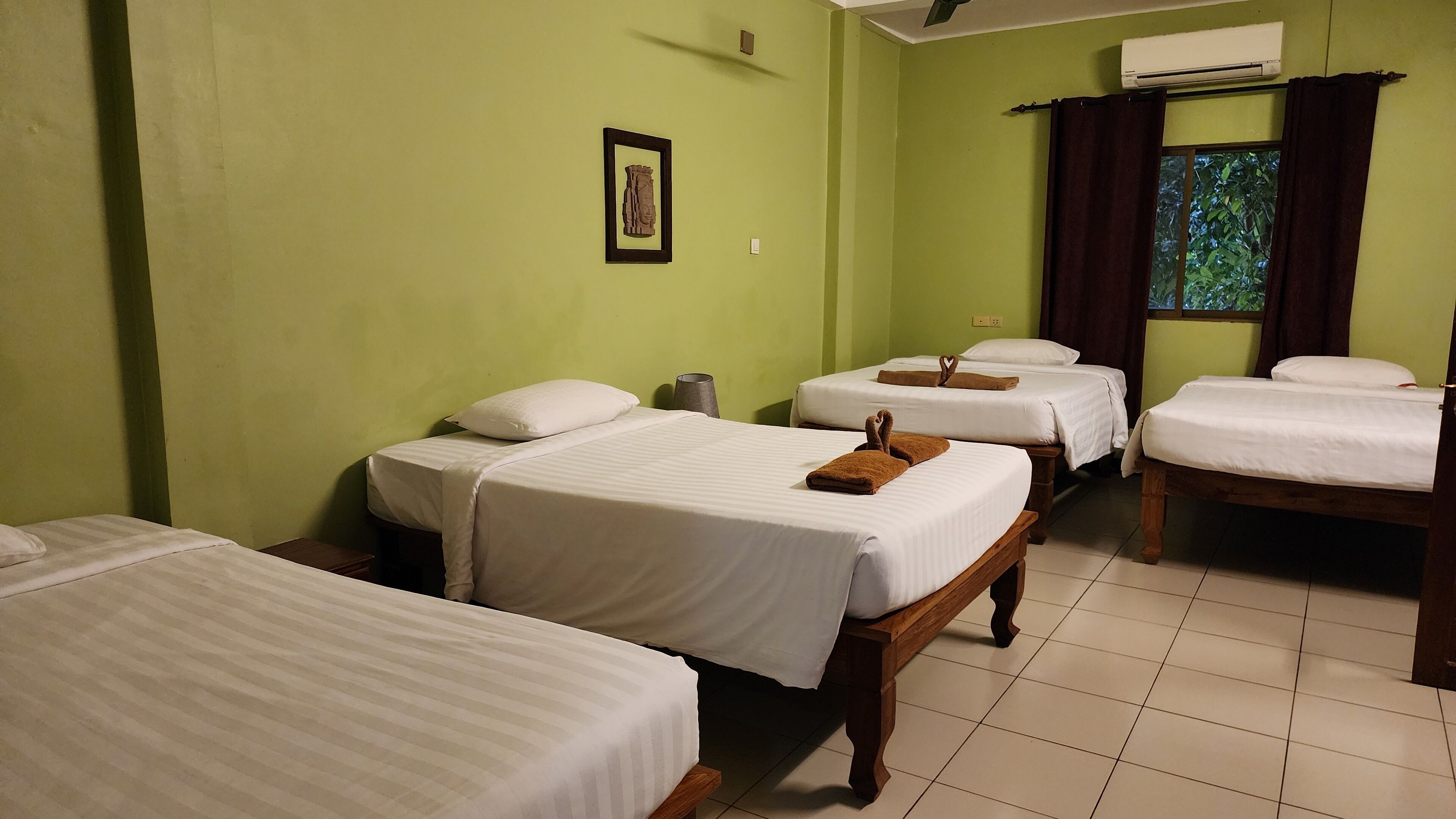 A hotel room with four single beds, a small table, a wall-mounted air conditioner, and a window with curtains.