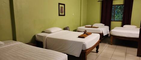 A hotel room with four single beds, a small table, a wall-mounted air conditioner, and a window with curtains.