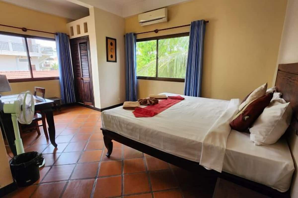 Een hotelkamer met een bed, een bureau, een stoel, een raam met gordijnen en een betegelde vloer.