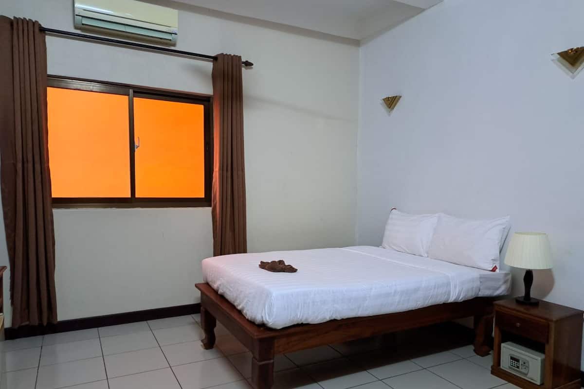 Een hotelkamer met een bed, een nachtkastje met lamp, een raam met gordijnen, en een luchtverkoeler aan de muur.