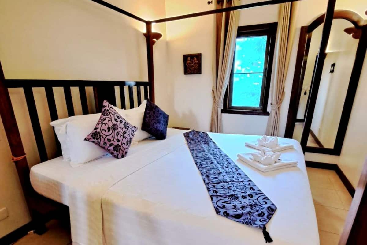 Een net opgemaakt bed in een hotelkamer, met een houten hoofdbord, een raam met gordijnen en een airconditioning op de muur.