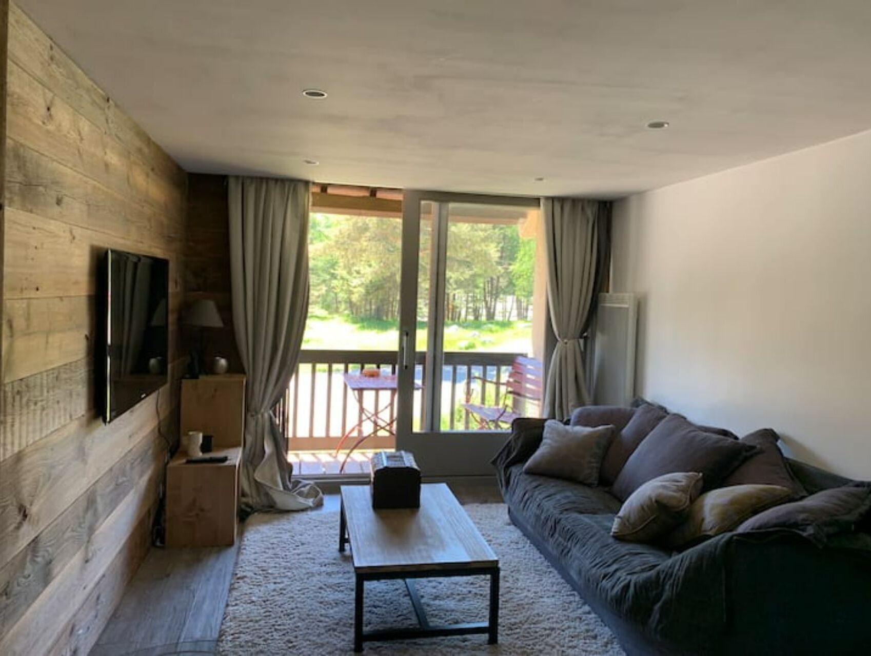 Une pièce chaleureuse avec un mur en bois, un téléviseur à écran plat, un canapé, une table basse et une vue sur une terrasse et la verdure environnante.
