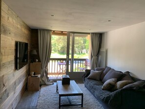 Quarto aconchegante com parede de madeira, TV de tela plana, sofá, mesa de centro e vista para um deck e vegetação lá fora.