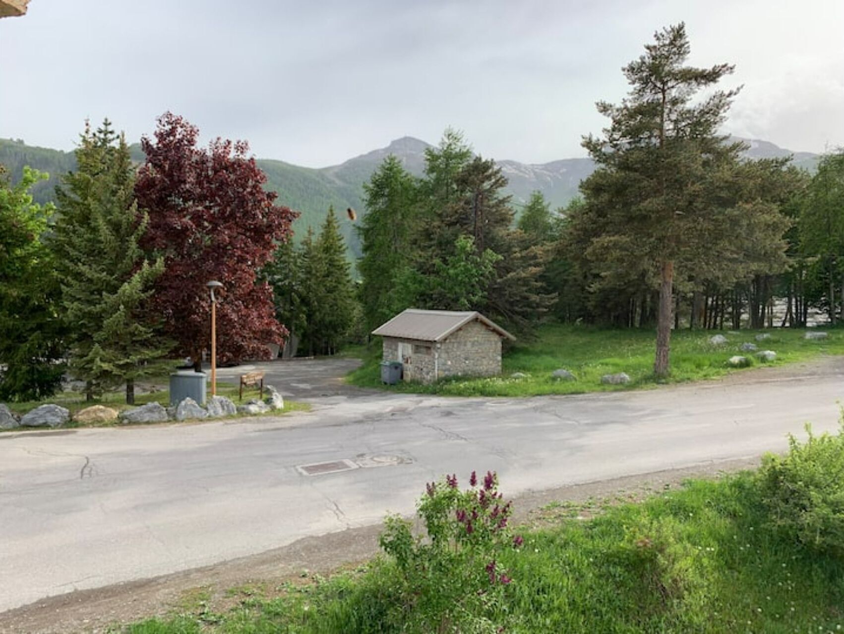 A rural road with a small stone building, surrounded by trees and mountains.