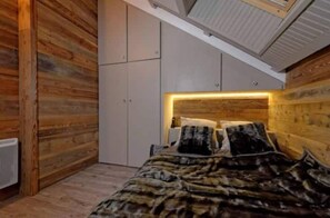 Quarto com teto inclinado, paredes de madeira, cama com colcha estampada e cabeceira com iluminação integrada.
