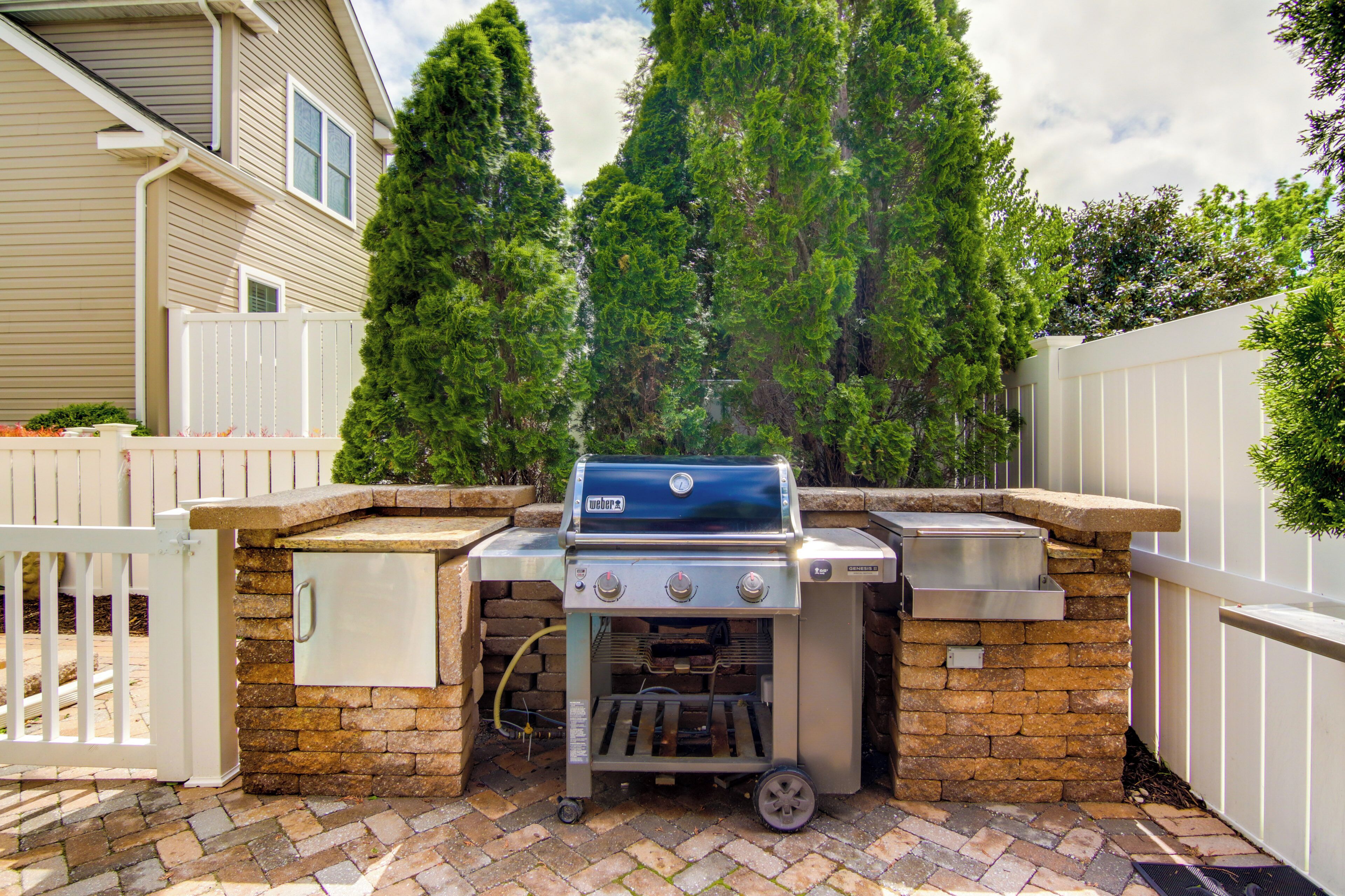 An outdoor barbecue grill with a stone base, a built-in sink, and a side cabinet, set against a brick patio with a white fence and tall trees in the background.