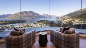 Una habitación moderna con un ventanal grande que ofrece una vista panorámica de montañas y un lago. Hay dos sofás a cuadros y una mesa redonda en el centro.