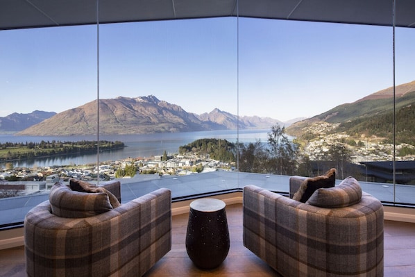 Una habitación moderna con un ventanal grande que ofrece una vista panorámica de montañas y un lago. Hay dos sofás a cuadros y una mesa redonda en el centro.