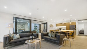 Een moderne woonkamer met een bank, salontafel en een eethoek. Achtergrond is een keuken met een koelkast en een eettafel.