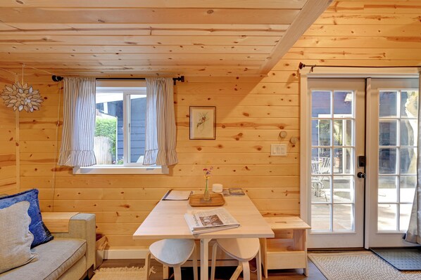 Interior de una cabaña de madera con una mesa de comedor, sillas, un sofá y una ventana con cortinas.