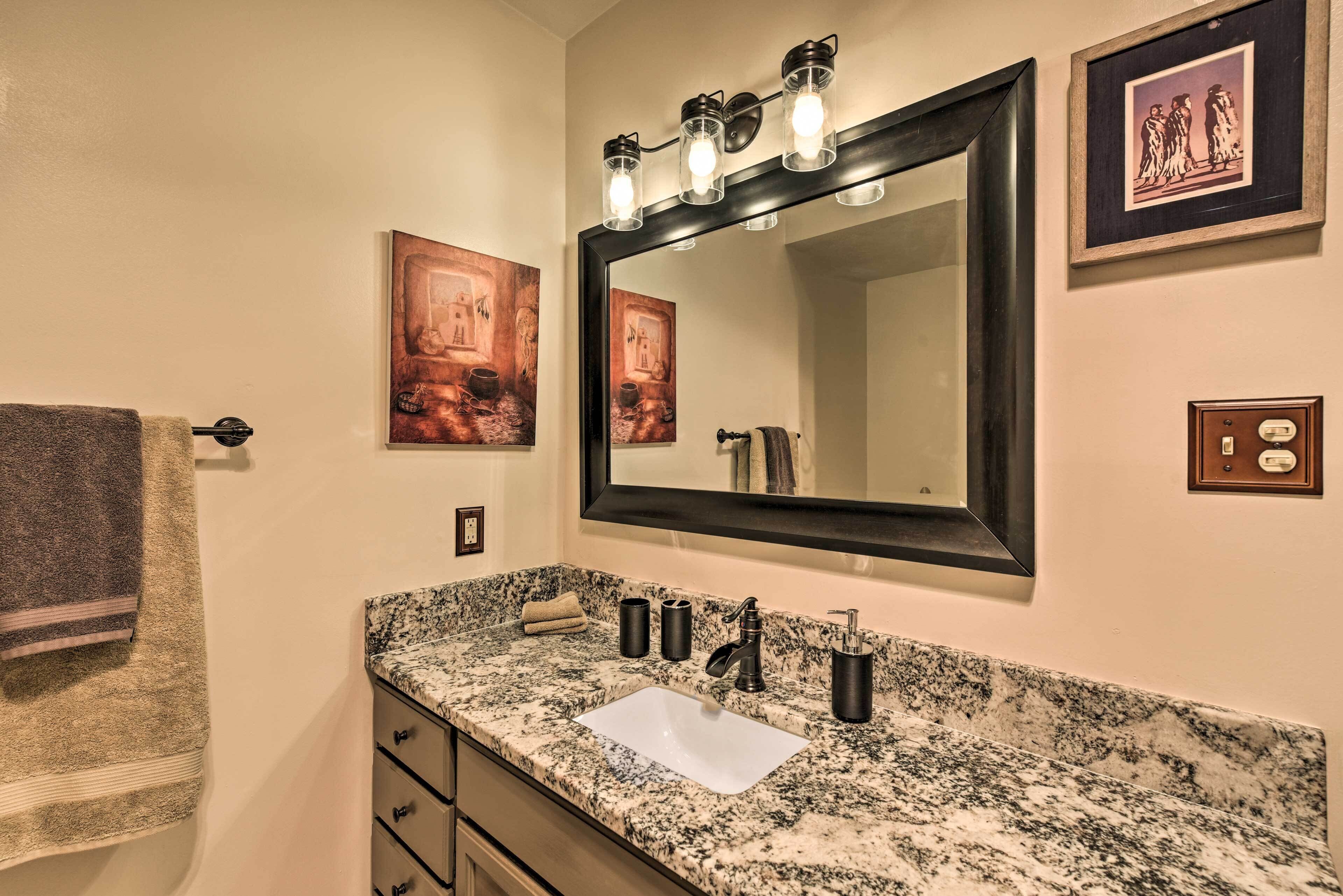 A bathroom with a granite countertop, a white sink, a large mirror with lights, and two framed pictures on the wall.