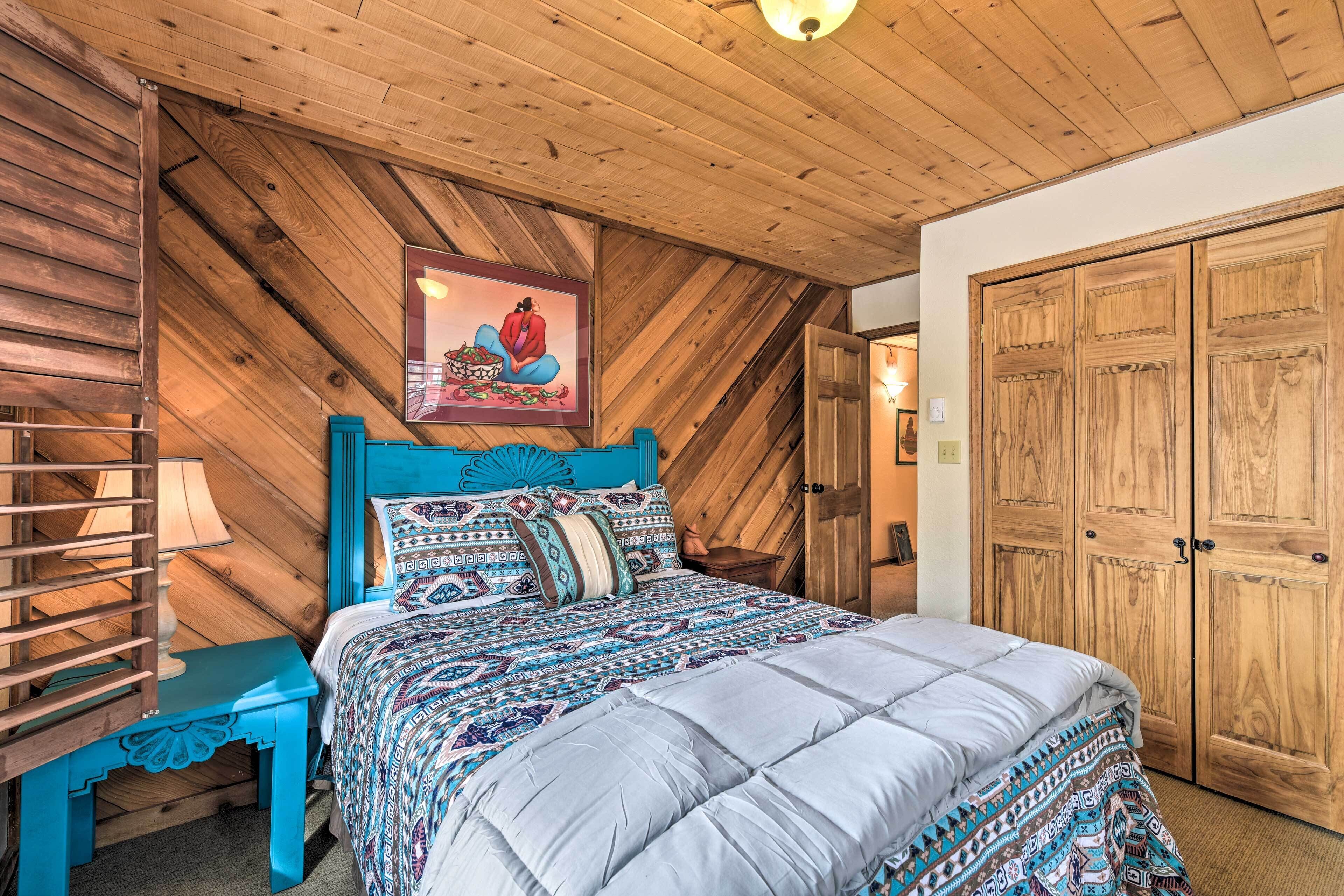 A bedroom with a wooden ceiling, a bed with patterned bedding, a blue nightstand, and a wooden wardrobe.