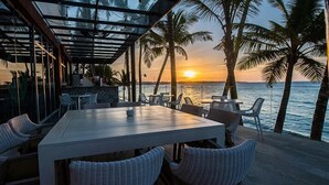 Zona pranzo all'aperto con tavolo bianco e sedie in vimini, affacciata su una spiaggia con palme e un tramonto.