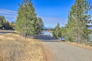 A paved road flanked by tall pine trees, leading to a body of water with a distant mountain range. - Woodsy Riverfront Retreat in Trout Creek Montana! (Trout Creek)