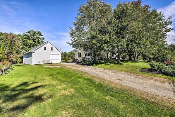 A white house with a front porch, a dirt driveway, and a large tree in front.