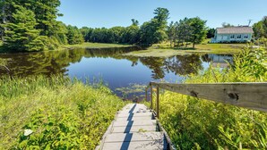 A wooden boardwalk leading to a calm lake surrounded by lush greenery and a house in the background. - On-site Pond: Spacious Family Home in Jefferson! (Jefferson)