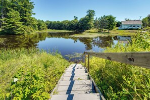 A wooden boardwalk leading to a calm lake surrounded by lush greenery and a house in the background. - On-site Pond: Spacious Family Home in Jefferson! (Jefferson)