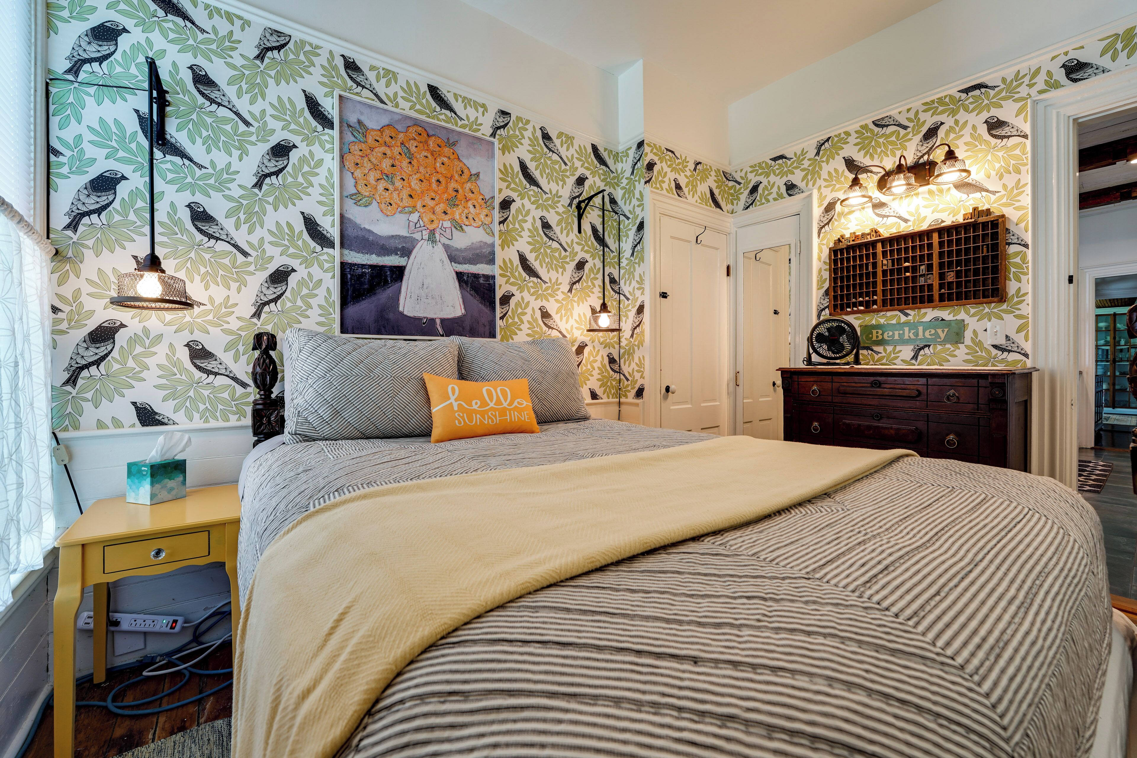 A bedroom with a bed, a bedside table, a nightstand, a wall with bird and leaf patterns, a painting, and a dresser with a mirror.