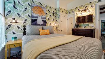 A bedroom with a bed, a bedside table, a nightstand, a wall with bird and leaf patterns, a painting, and a dresser with a mirror.