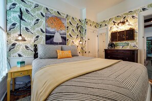 Un dormitorio con cama, mesita de noche, cómoda con espejo y una pared decorada con motivos de aves y hojas, además de un cuadro.
