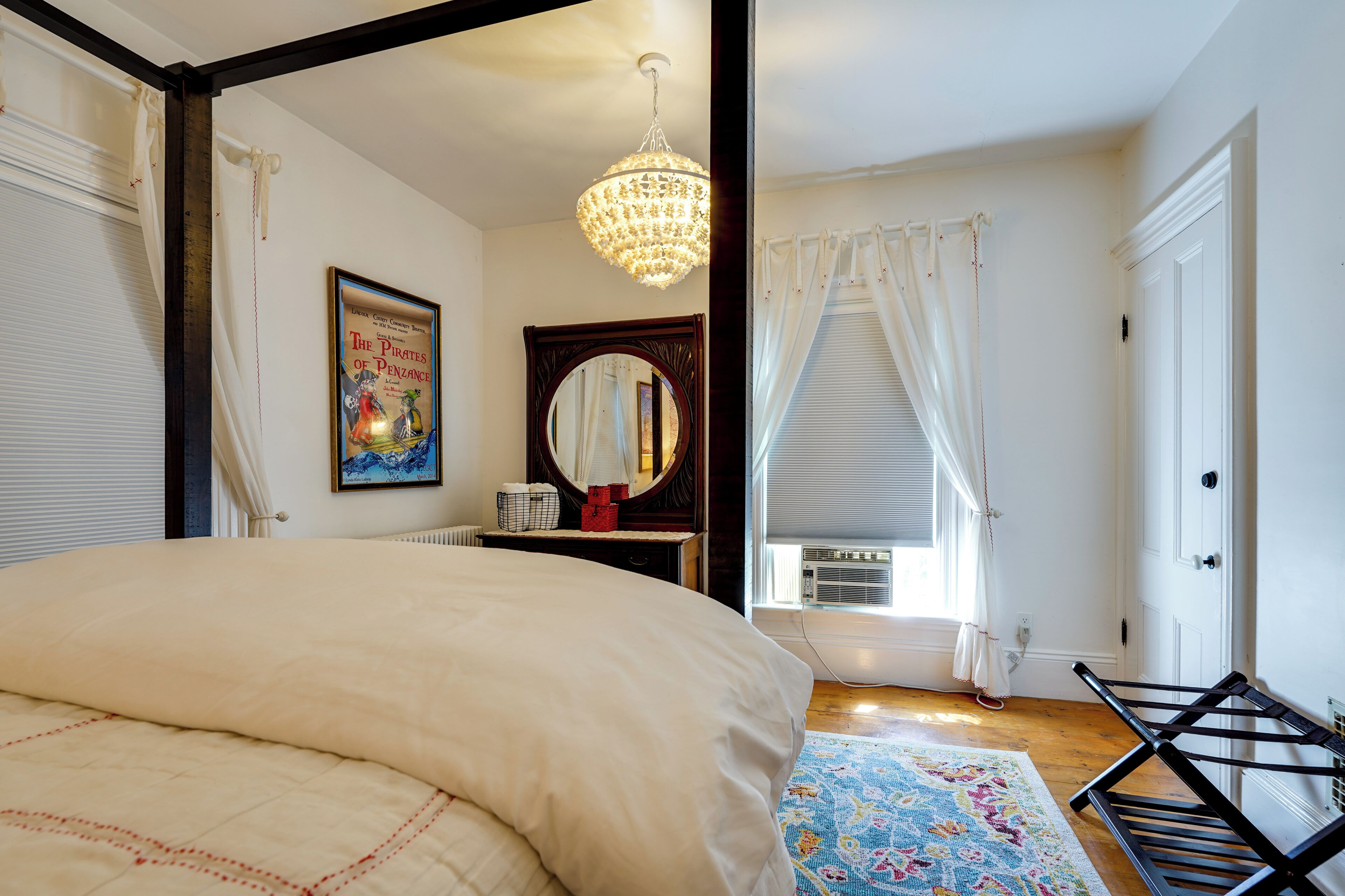 A bedroom with a large bed, a chandelier, a framed poster, a mirror, and a window with curtains.