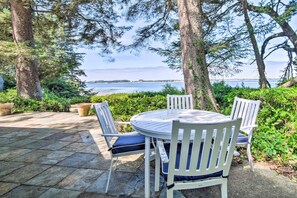 A round table with four white chairs and blue cushions, surrounded by stone tiles and greenery, overlooking a body of water.