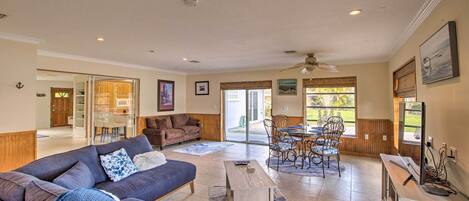 A spacious living area with a sofa, coffee table, and dining table. There is a door leading to another room, and a window with blinds.