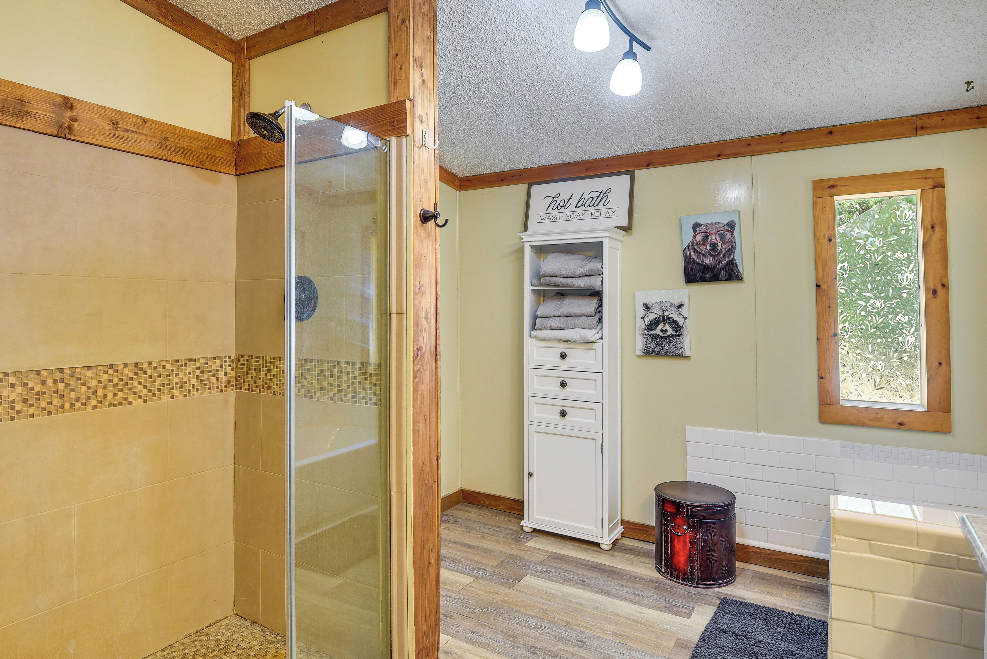 A bathroom with a glass shower enclosure, a white cabinet with towels, a wooden floor, and a window with a view.