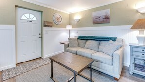 Ein Wohnzimmer mit einer weißen Tür, auf der „EXIT“ steht, einem beige farbenen Sofa, einem kleinen Tisch, einer Lampe und einer Wanduhr.