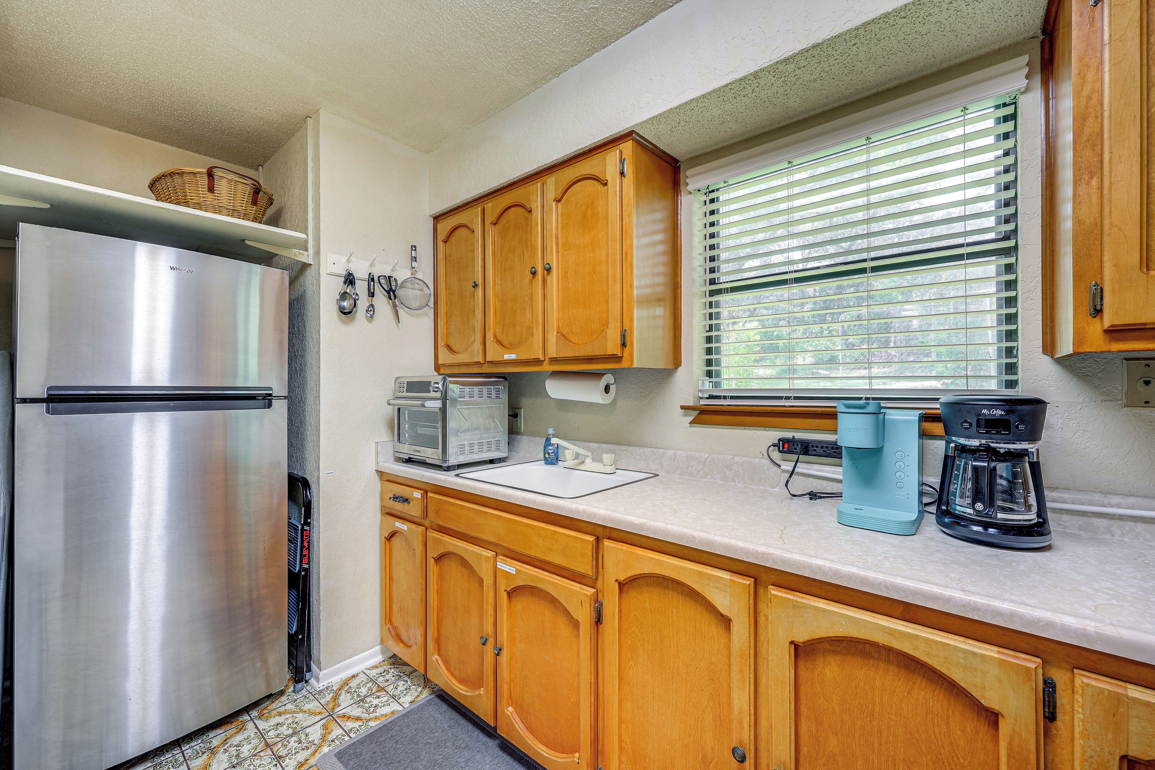 A kitchen with wooden cabinets, a stainless steel refrigerator, a microwave, a coffee maker, and a window with blinds.